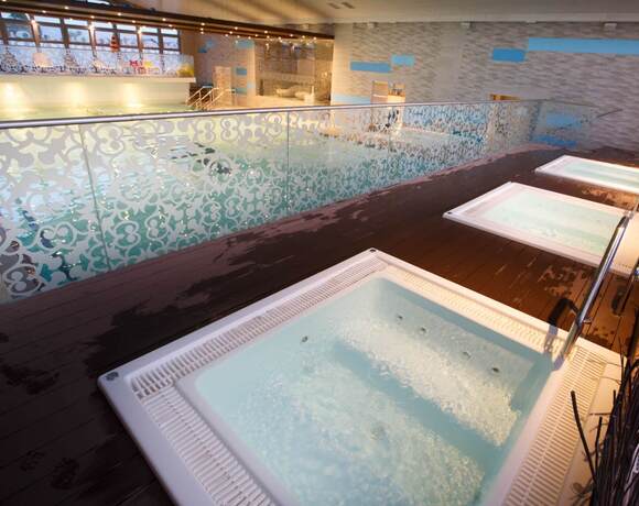 Three jacuzzis on a wooden terrace with a decorative glass balustrade, a view of the illuminated pool and an elegant, modern spa interior