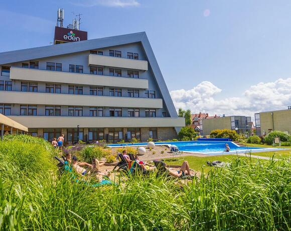 Hotel z skośnym dachem i tarasowymi balkonami, zewnętrzny basen, leżaki na trawniku i goście opalający się w pogodny dzień.