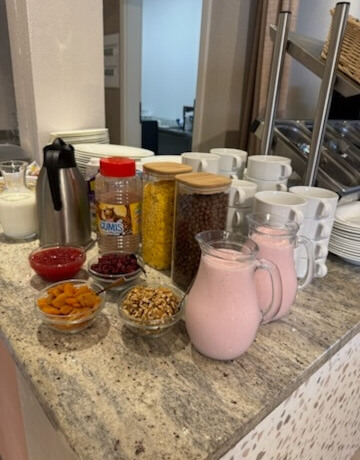Breakfast buffet: jugs of pink yogurt, jars of cereals and granola, dried fruit and nuts, a coffee thermos, stacks of cups and plates.