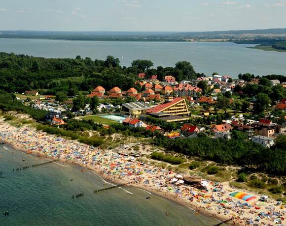 A sandy, crowded beach with umbrellas, a family resort by the dunes; a triangular hotel with a pool, a pine forest and a view of the bay