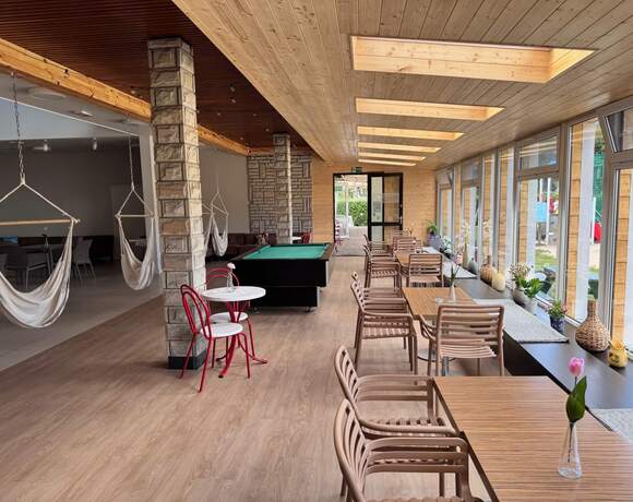 Cozy lounge with large windows, hammock chairs, tables, a pool table and a wooden ceiling with skylights.