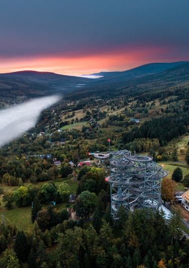 Sunset mountain valley with mist, forest trails and a spiraling treetop observation tower above countryside lodges.