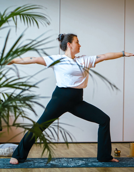 Guest practicing Warrior II yoga in a hotel wellness studio, leafy plants, yoga mat and relaxation props nearby.