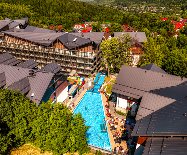 Outdoor pool with swimmers, water slides, sun loungers and balconies of a modern mountain hotel surrounded by forest.