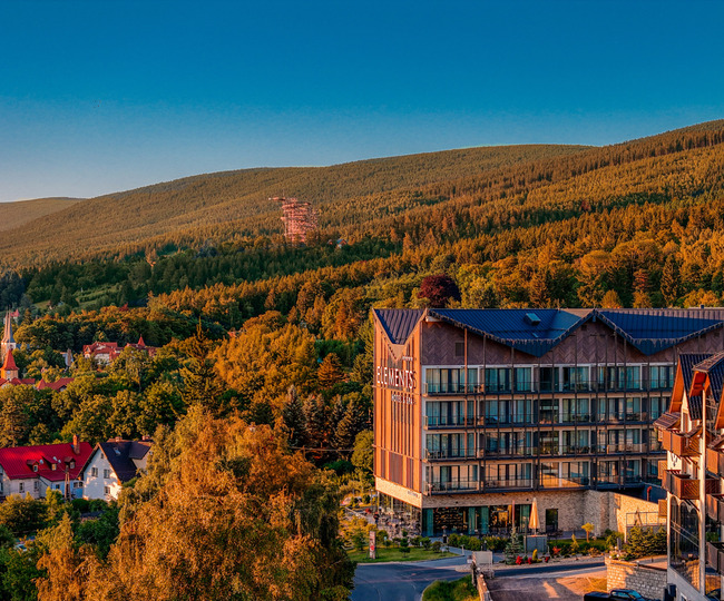 Modernes Elements Hotel & Spa mit Balkonen und Außenbereich, neben Dorfkirche und bewaldeten Bergen im Abendlicht.