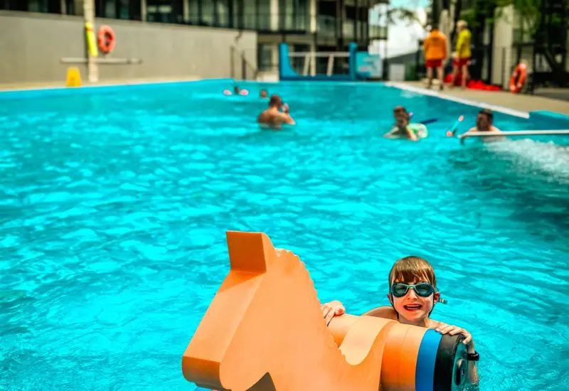 Hotel outdoor pool with a smiling child in goggles on a colorful horse-shaped float, modern lodge building and lifeguards nearby