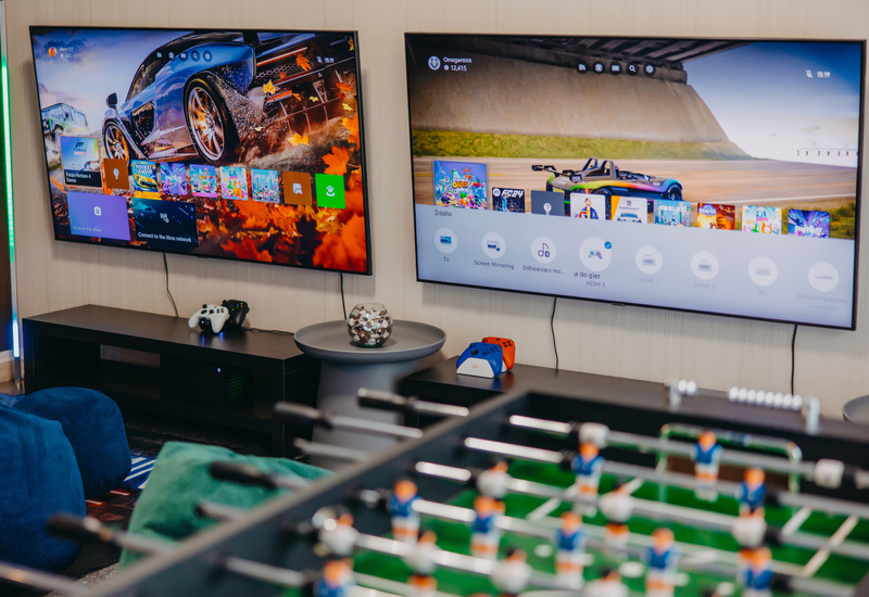 Gaming lounge with two wall-mounted TVs displaying racing games, consoles and controllers, foosball table and comfy seating.