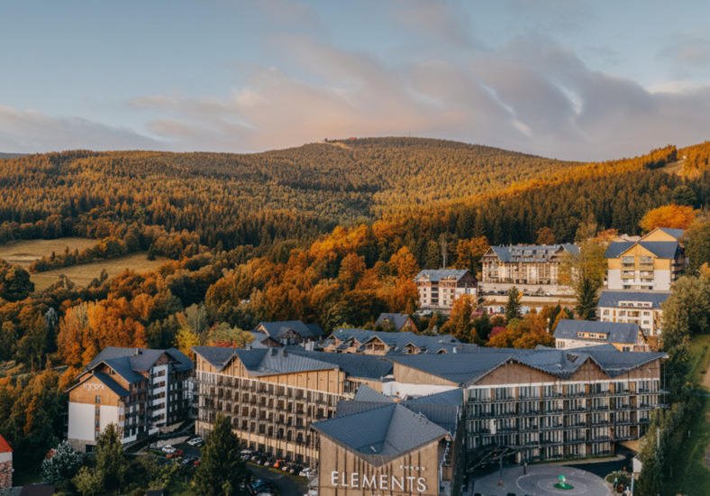 Elements mountain resort with timber-clad hotel buildings, terraces, parking and autumn forested hills at golden hour.