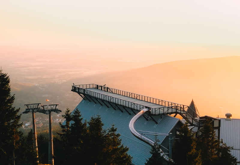 Rooftop observation deck with a winding alpine coaster track above the treeline, cable‑car towers and a golden sunrise over the valley