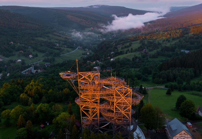Sunlit treetop observation walkway tower with spiral paths above forest, valley views, misty hills and scattered cottages at sunset.