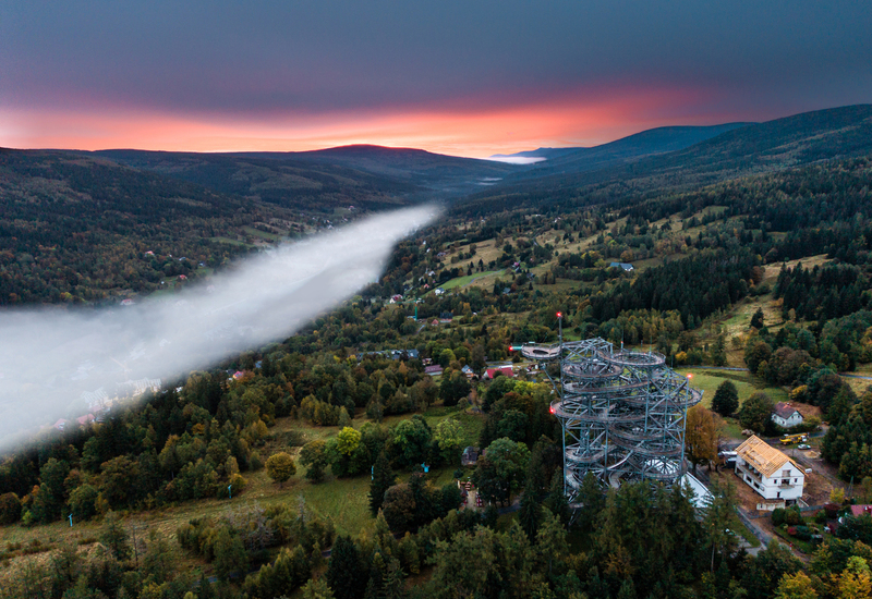 Sunset mountain valley with mist, forest trails and a spiraling treetop observation tower above countryside lodges.