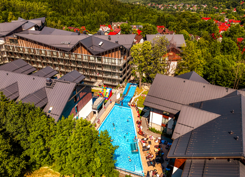 Outdoor pool with swimmers, water slides, sun loungers and balconies of a modern mountain hotel surrounded by forest.