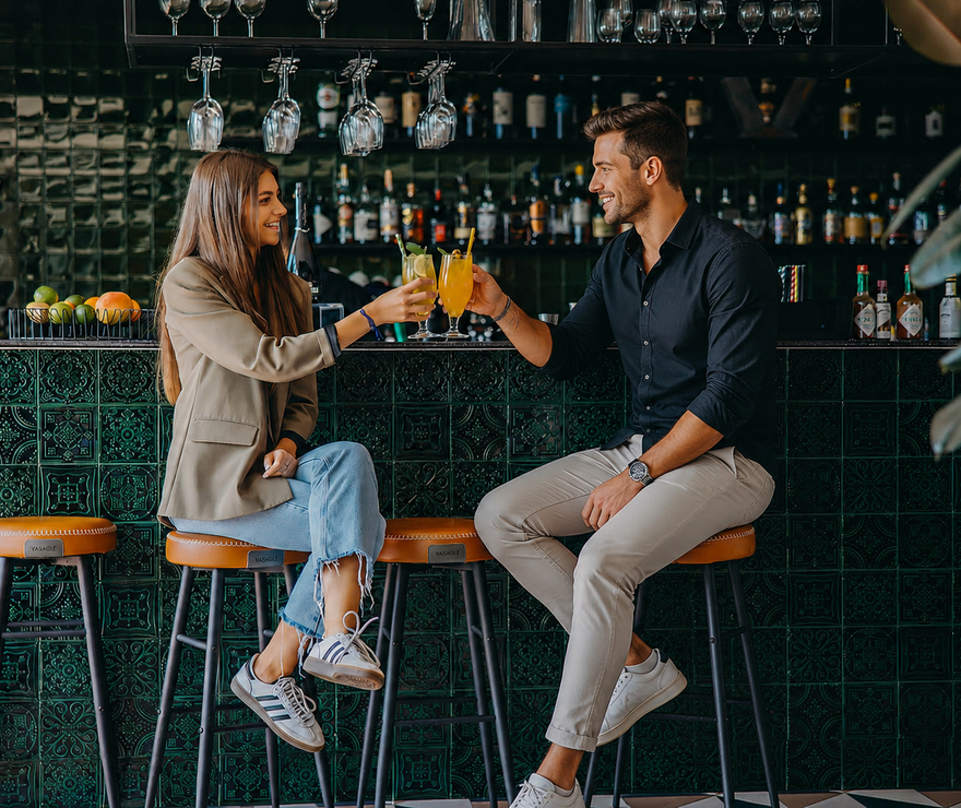 Para wznosi toast koktajlami przy stylowym barze hotelowym z zieloną mozaikową ladą, skórzanymi hokerami i wiszącymi kieliszkami
