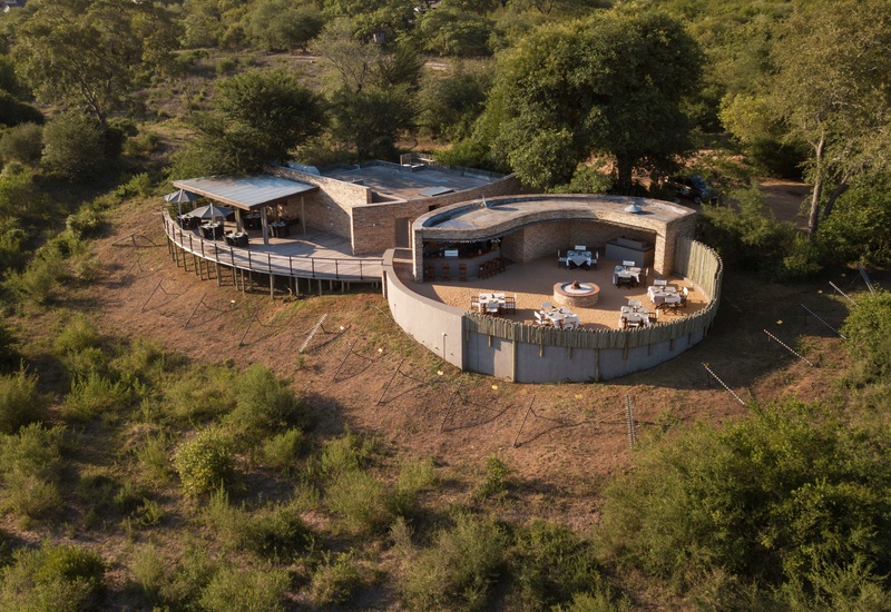 Curved stone safari lodge dining terrace with set tables, central fire pit, outdoor bar and covered raised deck overlooking bushveld.