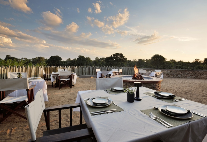 Safari lodge outdoor dining terrace with white-cloth tables, place settings and central fire pit overlooking tree-lined savanna at sunset