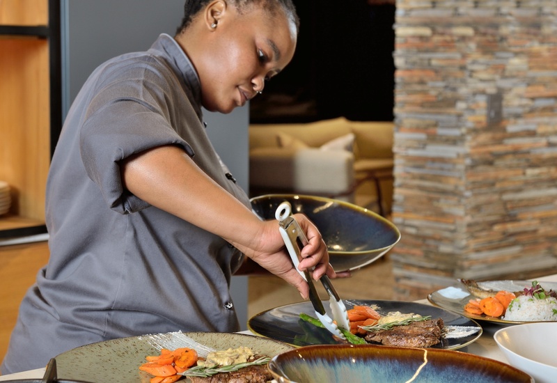 Chef plating steak and asparagus at an open-kitchen hotel station, ceramic bowls, roasted carrots and elegant stone-accent backdrop.