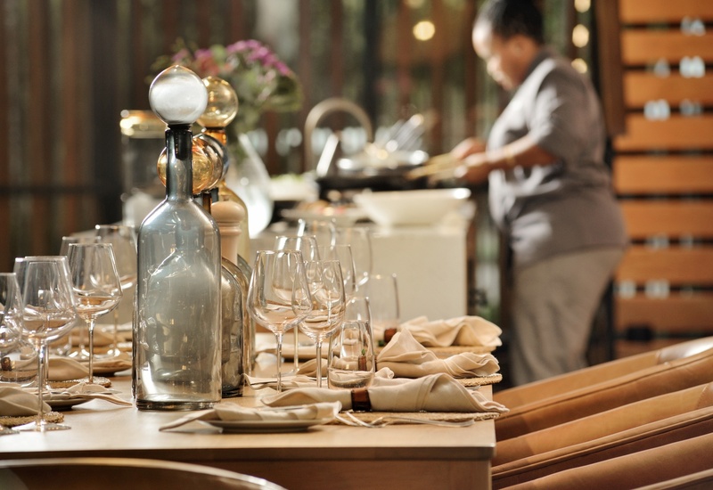 Elegant restaurant table set with wine glasses, glass carafes and folded napkins; server preparing dishes in background