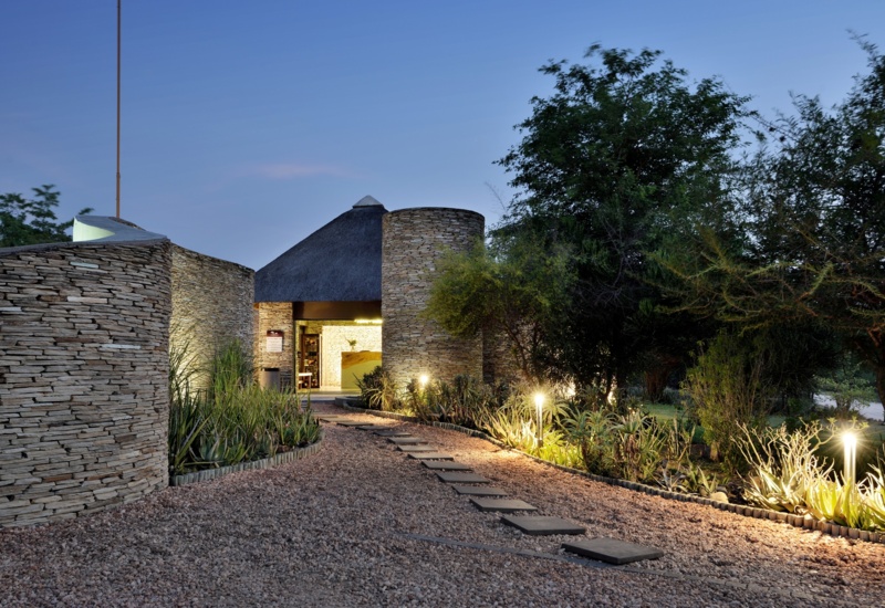 Thatched-roof stone lodge entrance with round stacked-stone walls, illuminated stepping-stone path and landscaped succulents to warm reception.