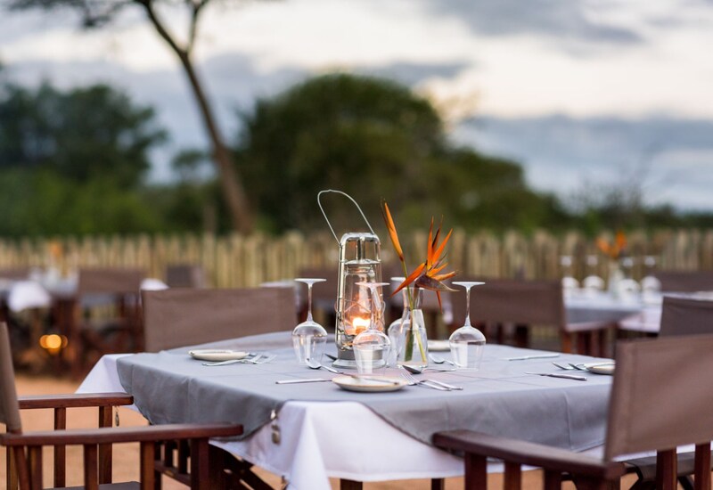 Outdoor hotel terrace dining at dusk with linen-covered table, lantern candle, bird-of-paradise flower and set wine glasses.