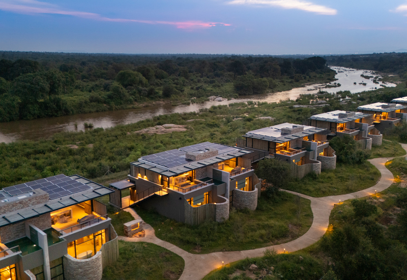 Riverside luxury villas with private terraces, solar roofs and lit winding paths overlooking a lush river and sunset-dusk savanna.