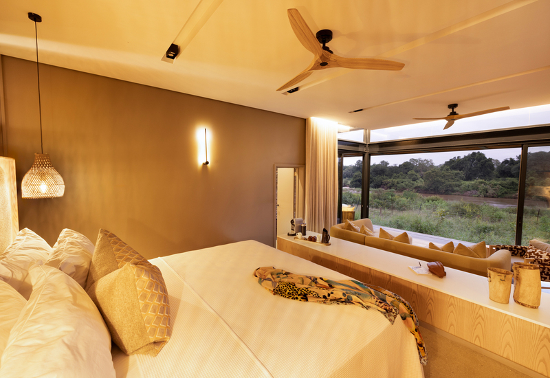 Luxury bedroom with king bed, woven pendant light, sofa seating and floor-to-ceiling glass opening to riverside view.