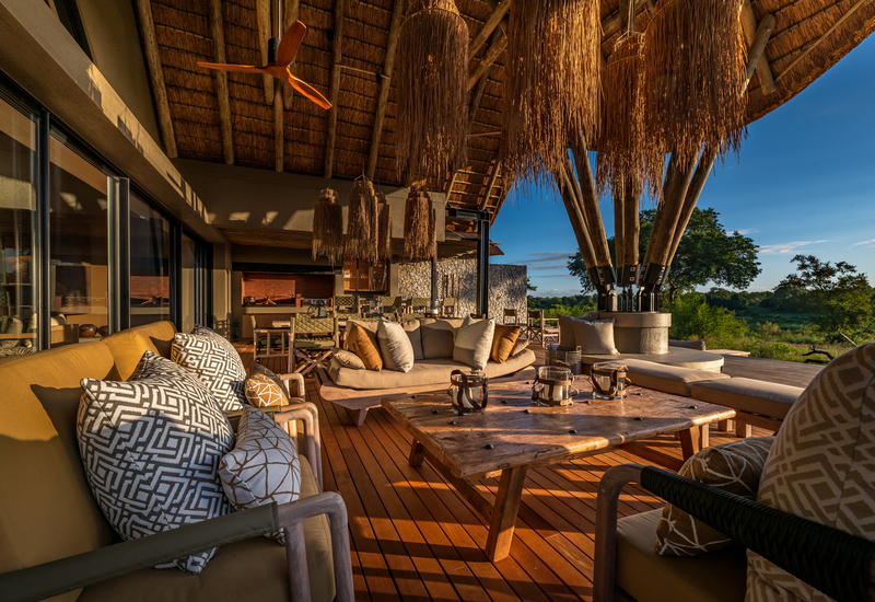 Thatched-roof outdoor lounge with plush sofas and patterned cushions, rustic wooden coffee table, straw pendant lights and treetop sunset views.