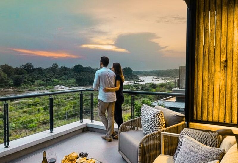 Couple on a private balcony at sunset overlooking a river and green bushland, cozy outdoor lounge with wine and snacks on the table.