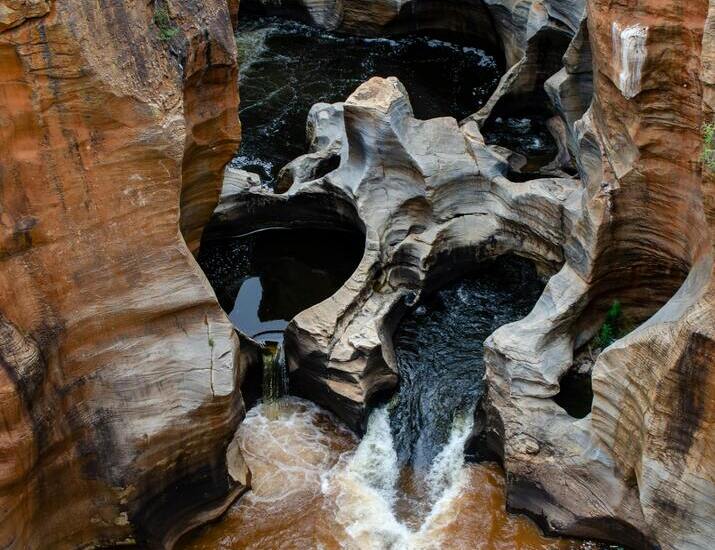Dramatic carved rock canyon with swirling potholes and rust-colored river water cascading through narrow rock channels.