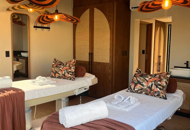 Twin spa massage tables with rolled towels, slippers and patterned pillows under warm sculptural pendant lights, wood accents