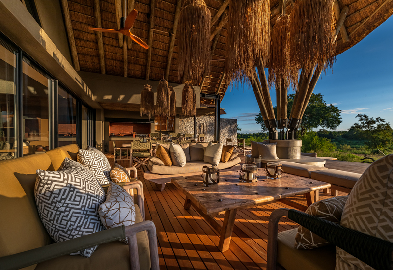 Thatched-roof outdoor lounge with plush sofas, patterned pillows, rustic wooden table and hanging reed lights overlooking treed savannah