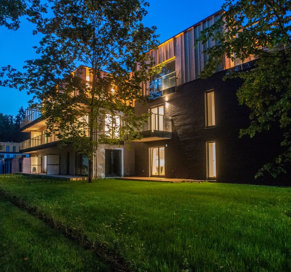 Nowoczesny butikowy hotel o zmierzchu z oświetlonymi balkonami, dużymi oknami, drewnianymi detalami i trawiastym tarasem wśród drzew