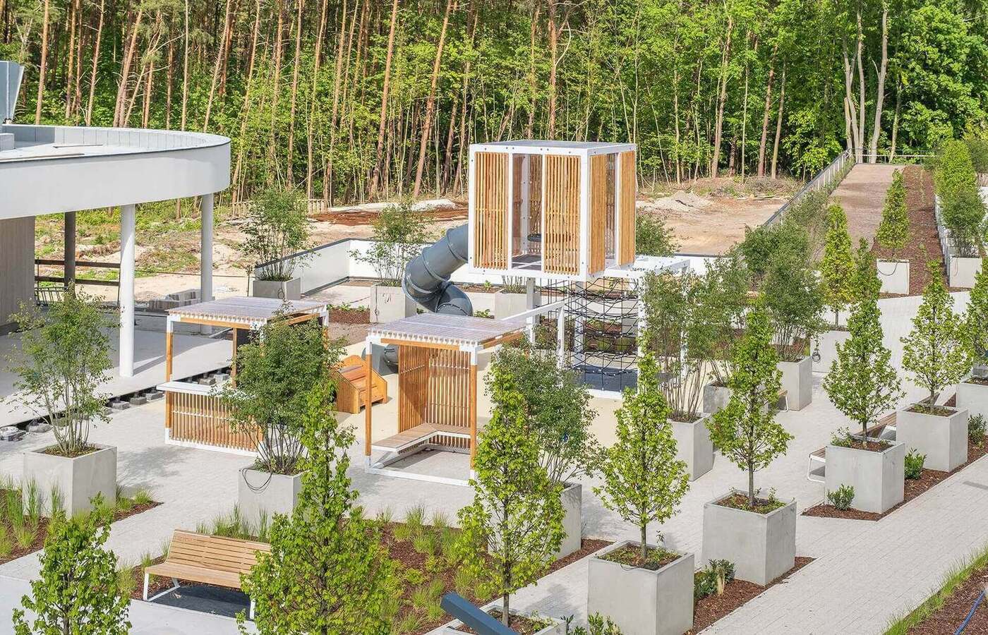 Nowoczesny taras z drewnianymi, zacienionymi kabinami, ławkami, nasadzeniami i podwyższoną wieżą zabaw z rurkową zjeżdżalnią, las w tle.