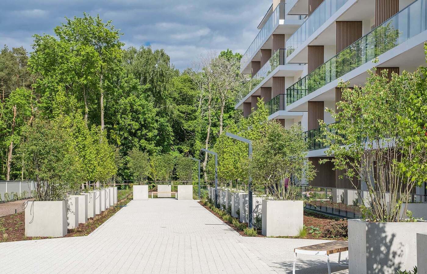 Nowoczesny hotel z przeszklonymi balkonami przy zadbanej promenadzie z betonowymi donicami, ławeczkami i widokiem na las