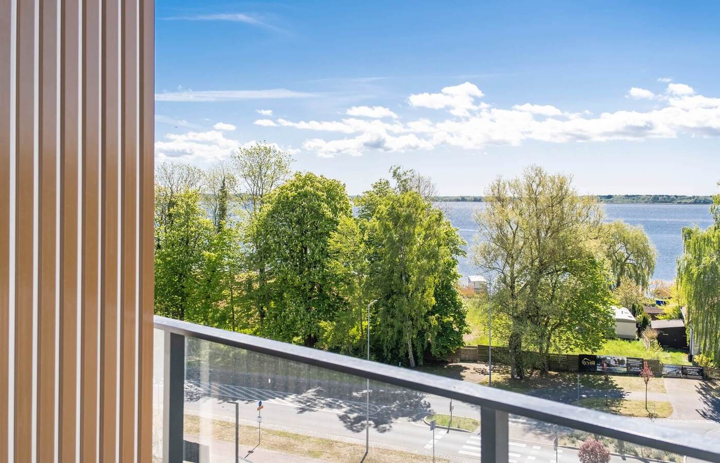 Balkon ze szklaną balustradą i pionową drewnianą okładziną, widok na zielone drzewa i spokojne jezioro pod jasnym niebem