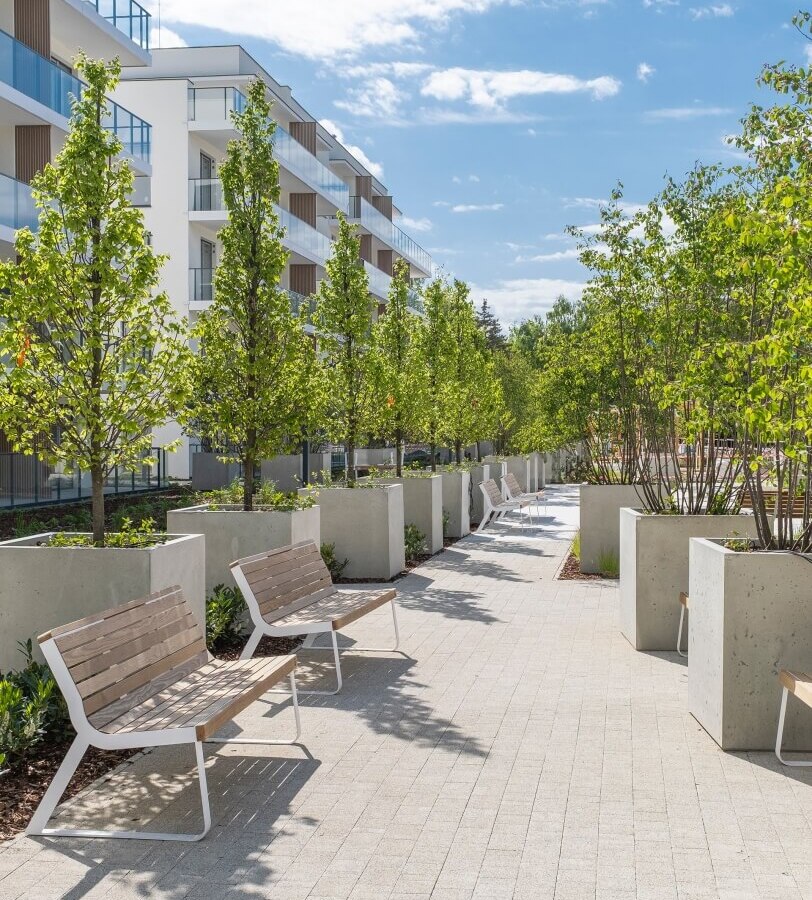 Słoneczny dziedziniec hotelowy z drewnianymi ławkami, dużymi betonowymi donicami, młodymi drzewami i pokojami z przeszklonymi balkonami