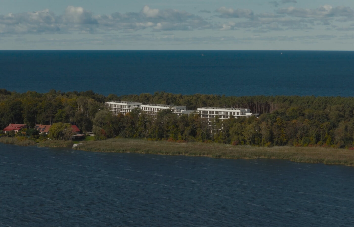 Biały hotel niskiej zabudowy z balkonami w sosnowym lesie, z widokiem na morze i spokojną lagunę — zaciszny wypoczynek