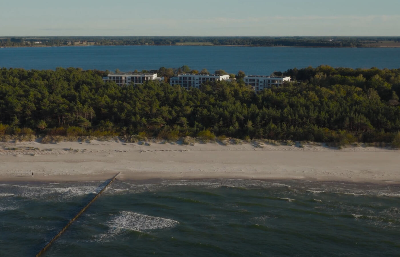 Nowoczesne niskie budynki hotelowe za sosnowym lasem, piaszczysta plaża z drewnianym falochronem i widokiem na morze, spokojny wypoczynek