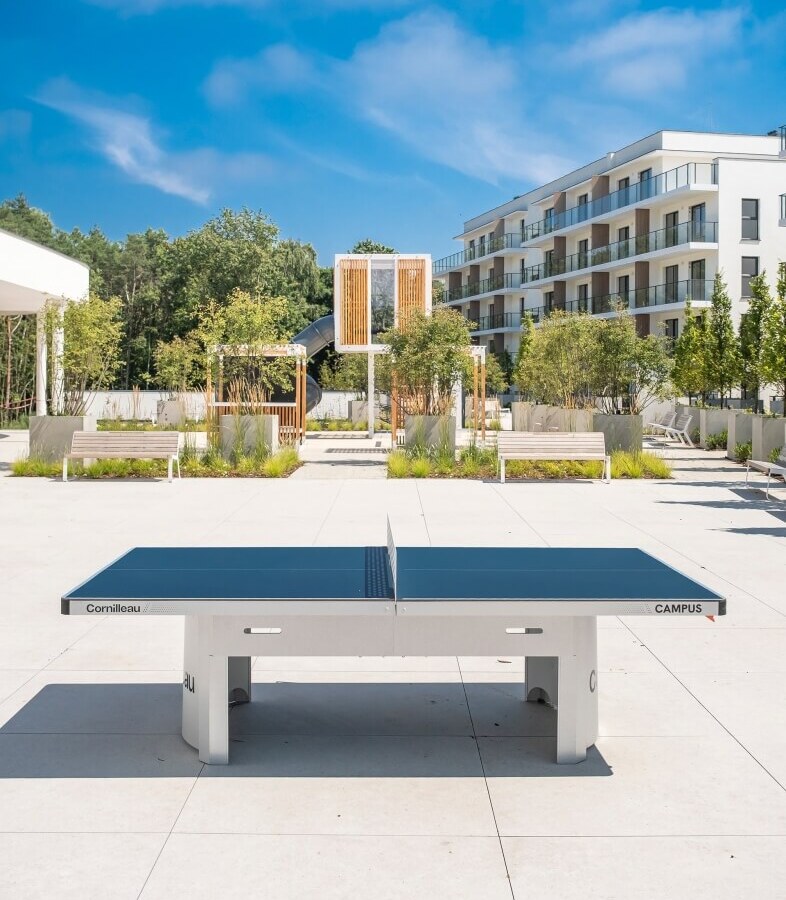 Stół do ping‑ponga na słonecznym dziedzińcu hotelu, nowoczesny budynek z balkonami, ławki i zadbana zieleń.