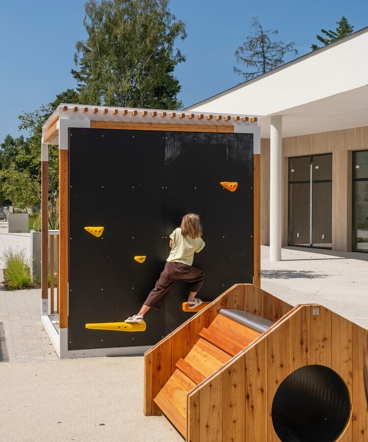 Zewnętrzny plac zabaw dla rodzin, dziecko na czarnej ściance wspinaczkowej z żółtymi chwytami i drewnianym tunelem przy nowoczesnym hotelu