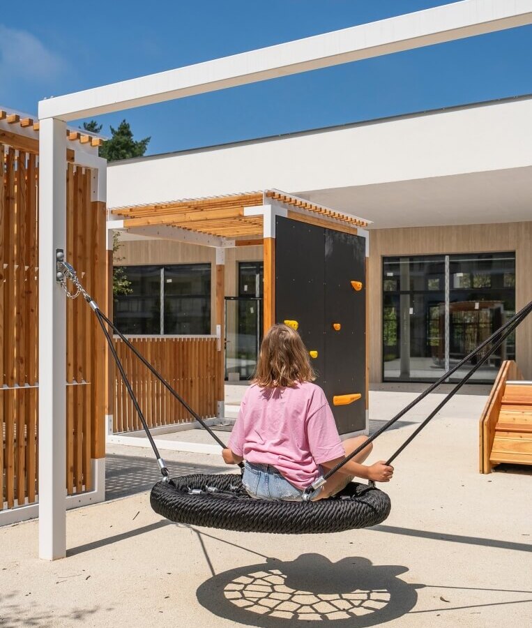 Młody gość na okrągłej huśtawce linowej na słonecznym patio hotelowym, pergole, ścianka wspinaczkowa i miejsca do siedzenia