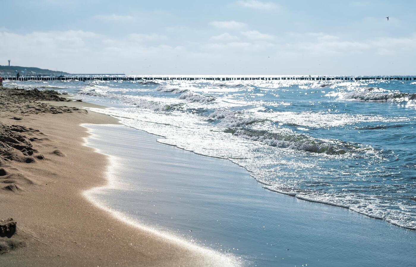 Piaszczysta plaża z delikatnymi falami i błyszczącym morzem, w oddali drewniany pomost — spokojne miejsce na spacery i relaks