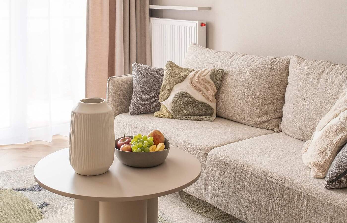 Strefa wypoczynkowa w apartamencie: beżowa sofa z dekoracyjnymi poduszkami, okrągły stolik z wazonem i miską owoców, lekkie zasłony i ciepłe lampy.