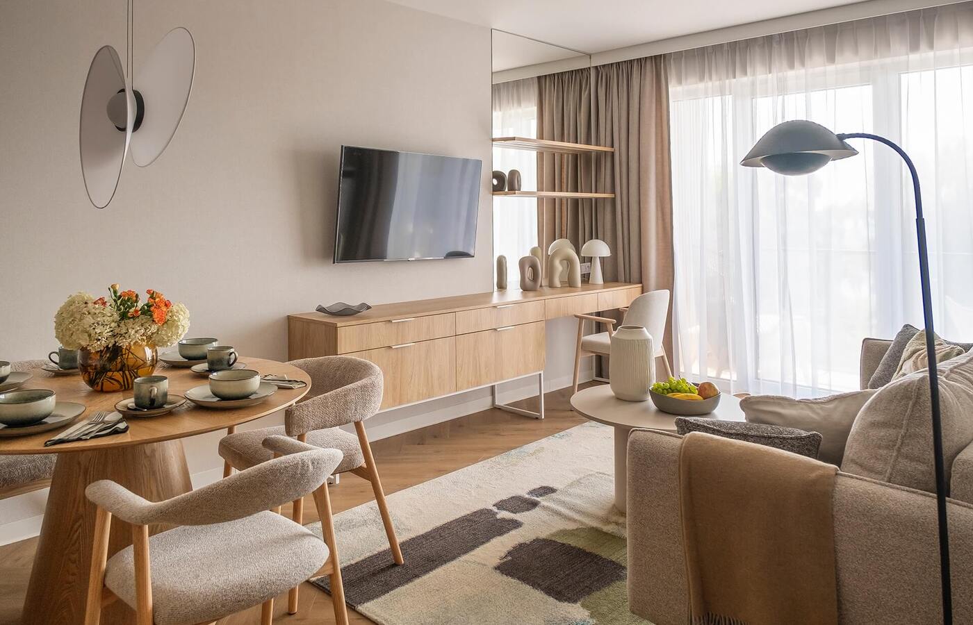 Jasny, nowoczesny salon w apartamencie z nakrytym stołem i wazonem kwiatów, TV na ścianie, drewniana komoda, firany i przytulna sofa.