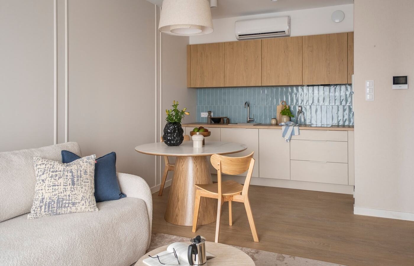 Jasne studio z przytulną sofą, okrągłym drewnianym stołem, aneksem kuchennym z dębowymi szafkami, błękitnym połyskującym panelem i klimatyzacją