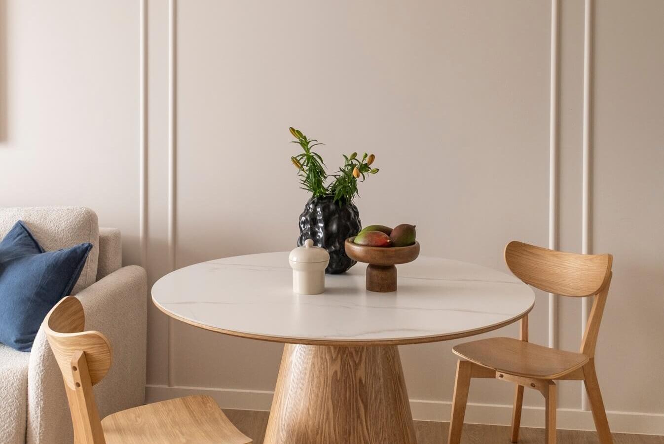 Przytulny kącik jadalny z okrągłym marmurowym stołem na drewnianej podstawie, dwoma dębowymi krzesłami, lampą i miską owoców.