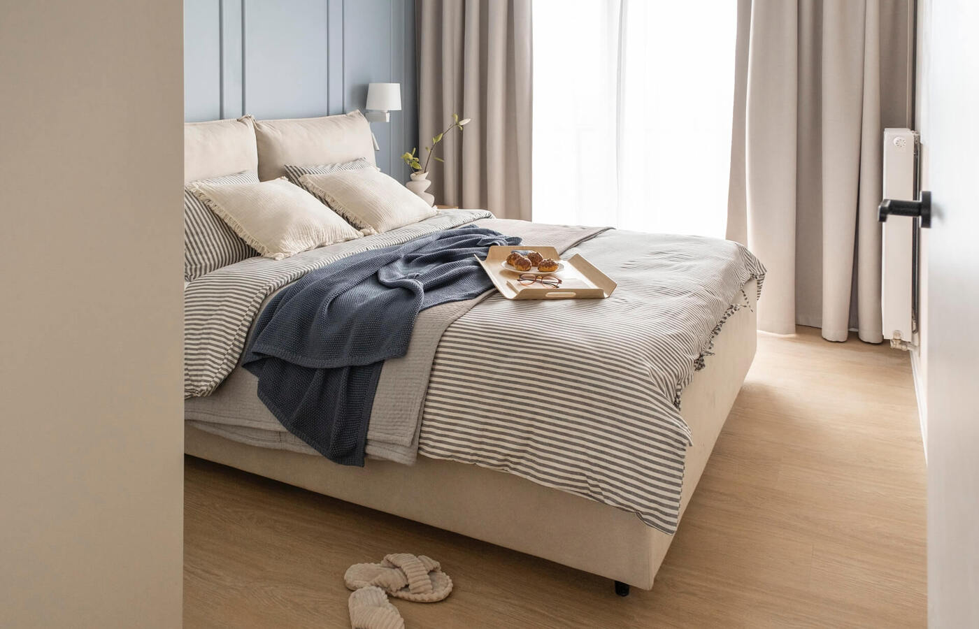 Jasny nowoczesny pokój hotelowy z pasiastą pościelą, miękkimi poduszkami, tacą z ciastkami na łóżku, lampką nocną i kapciami.