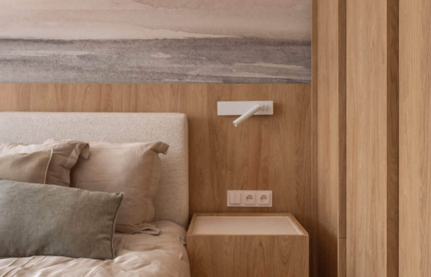 Róg łóżka z stonowaną pościelą, tapicerowanym zagłówkiem, drewnianą szafką nocną, lampką ścienną i pastelowym muralem morza