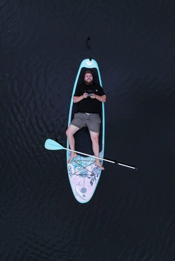 Gość relaksuje się na turkusowej desce SUP na spokojnej wodzie, boso i uśmiechnięty, wiosło poprzecznie