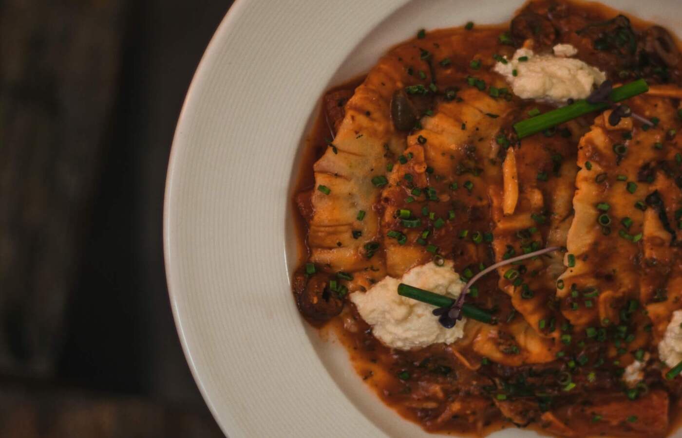 Domowe ravioli w sosie grzybowo-pomidorowym, kleksy ricotty i szczypiorek na białym talerzu na drewnianym stole