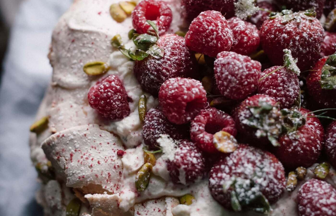 Beza Pavlova z bitą śmietaną, świeżymi malinami, pistacjami i posypana cukrem pudrem.